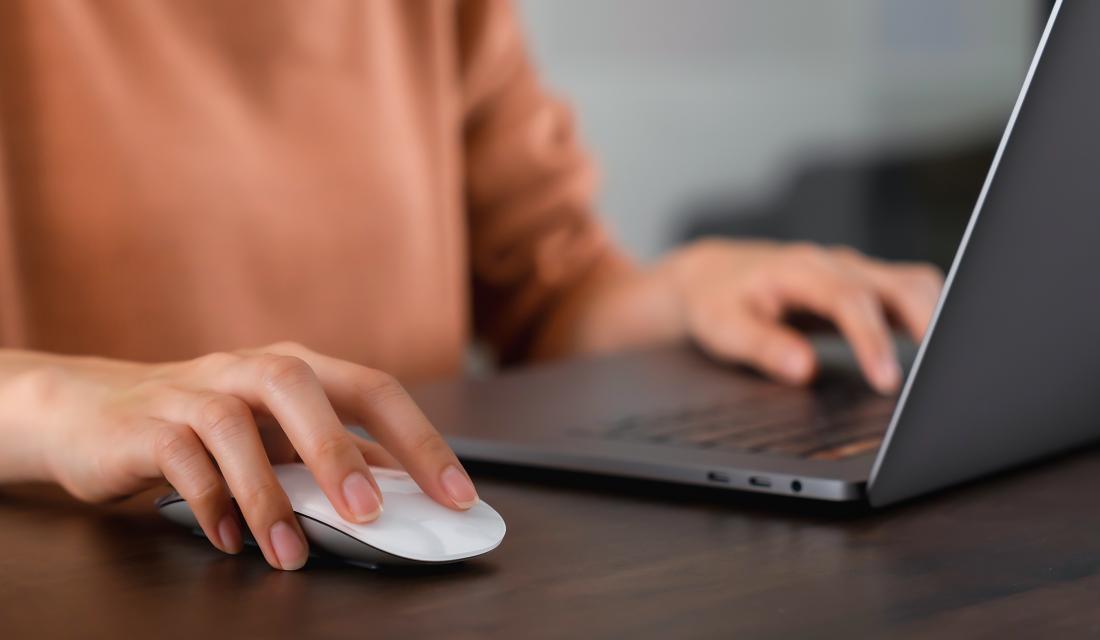 Eine Frau sitzt an ihrem Notebook und hält ihre Hand über eine Computermaus.