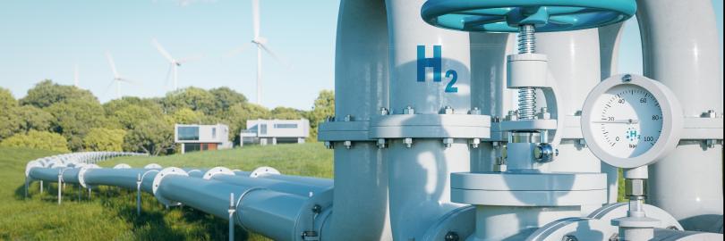 Eine Wasserstoff-Pipeline führt über eine grüne Wiese zu zwei Häusern. Im Hintergrund sind Windkraftanlagen zu sehen.