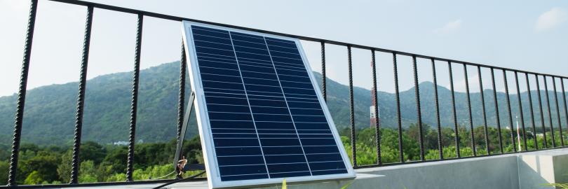 Auf einer sonnigen Dachterrasse lehnt ein einzelnes Solarmodul leicht schräg an einem schwarzen Metallgeländer. Im Hintergrund sind bewaldete Hügel und ein klarer Himmel zu sehen. Das Panel ist über ein Kabel angeschlossen, das über die niedrige Mauer der Terrasse führt. Vorne im Bild ragen einige grüne Zierpflanzen ins Bild und setzen einen natürlichen Akzent zu der technischen Anlage.