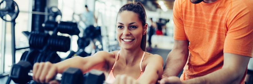 Frau trainiert mit Hanteln im Fitnessstudio, ein Trainer unterstützt sie dabei.