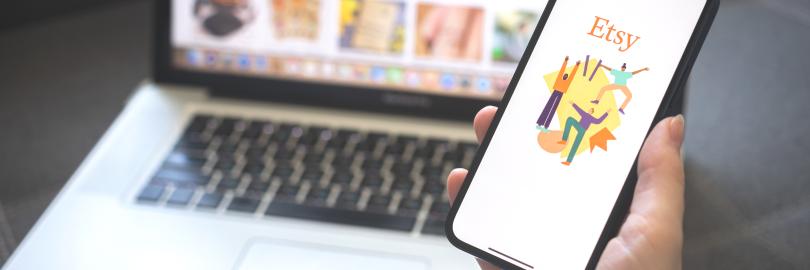 Eine Hand hält ein Handy, zu sehen die Etsy-App, im Hintergrund ein Laptop mit dem Etsy-Online Shop