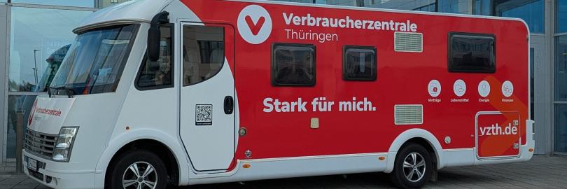 Ein großes, rot-weiß gestaltetes Infomobil der Verbraucherzentrale Thüringen steht vor einem modernen Gebäude mit Glasfassade. Auf der Seite des Fahrzeugs sind das Logo, der Schriftzug ‚Verbraucherzentrale Thüringen‘ sowie der Slogan ‚Stark für mich.‘ zu sehen. Außerdem sind Symbole für Themen wie Verträge, Lebensmittel, Energie und Finanzen abgebildet.