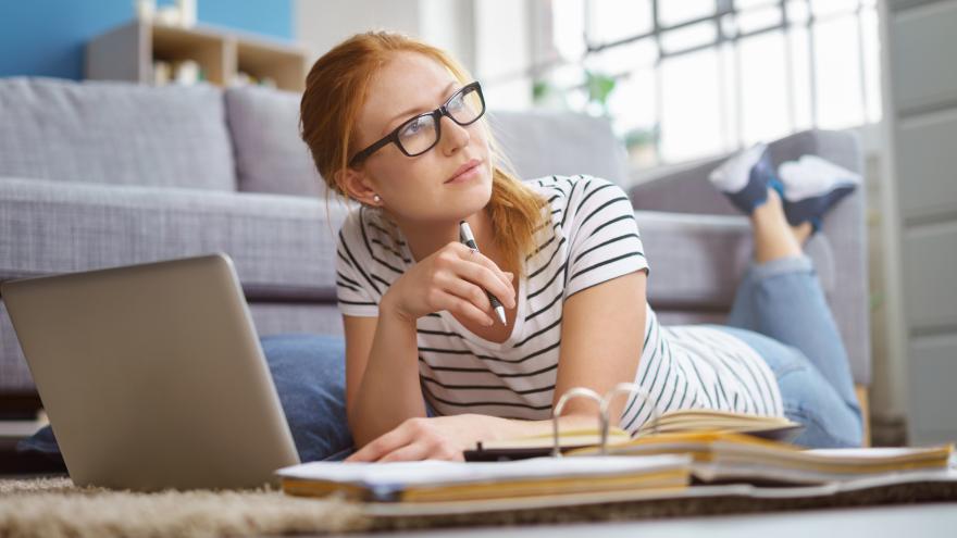 Eine junge Frau liegt in ihrem Wohnzimmer auf den Boden und arbeitet mit Laptop und Aktenordner.