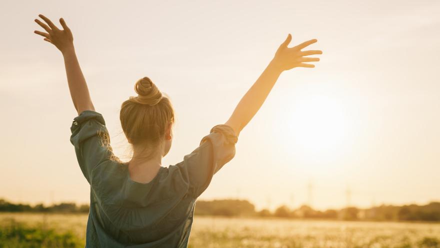 Eine junge Frau steht vor einem Getreidefeld in der Sonne und streckt die Arme in die Höhe.