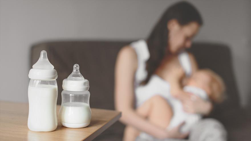 Zwei Babyflaschen auf einem Tisch, Mutter stillt Baby im Hintergrund