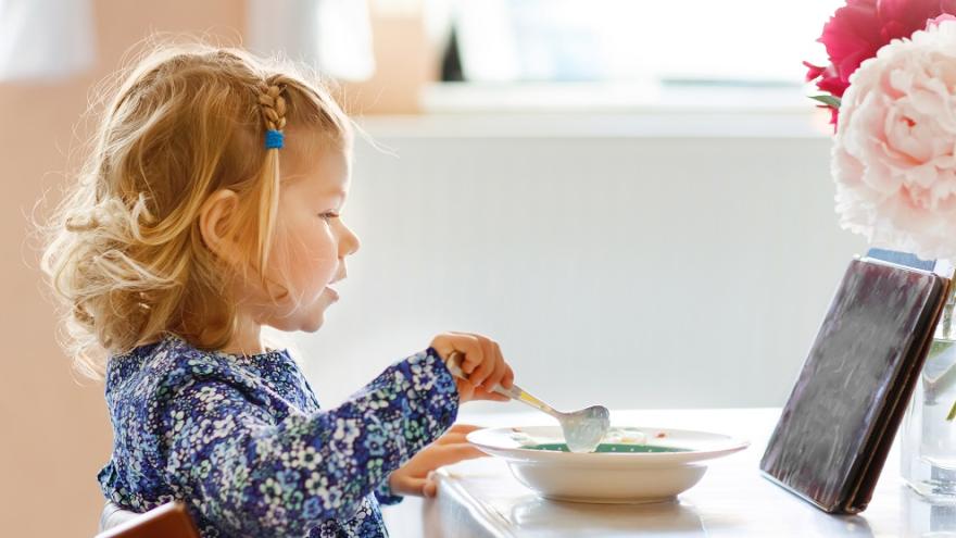 Ein Kind frühstückt während es auf einem Tablet etwas schaut.