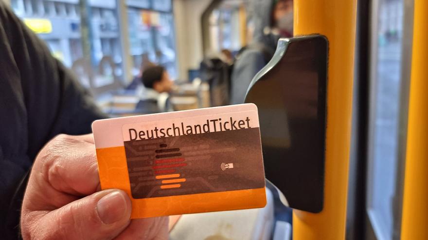 Eine Hand hält ein Deutschlandticket neben einen Stopp-Knopf an der Tür einer Straßenbahn