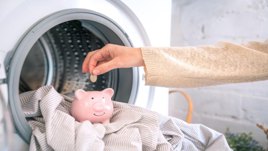 Eine Hand steckt ein Geldstück in ein Sparschwein, dass in einer Waschmaschine steht.
