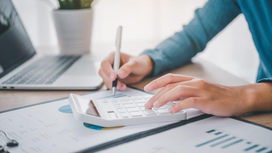 Großaufnahme eines Menschen am Schreibtisch mit Taschenrechner und Finanzunterlagen, im Hintergrund ein Laptop. 