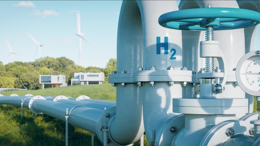 Eine Wasserstoff-Pipeline führt über eine grüne Wiese zu zwei Häusern. Im Hintergrund sind Windkraftanlagen zu sehen.