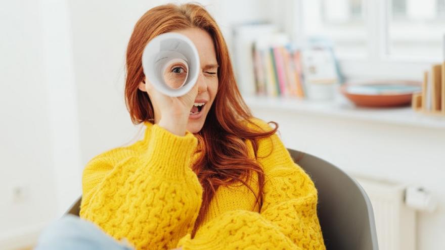 Eine rothaarige Frau in einem gelben Pullover schaut durch gerolltes Papier wie durch ein Fernrohr.