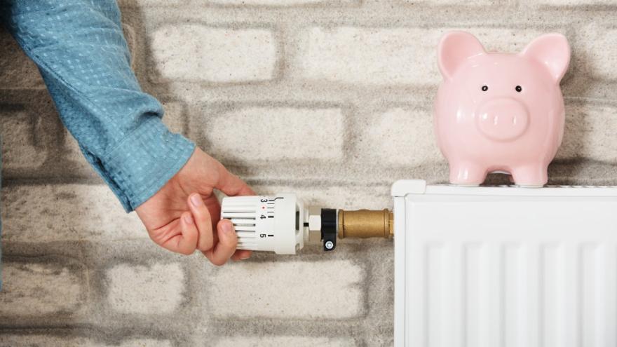 Eine Hand dreht an einem Heizungsthermostat, auf dem Heizkörper steht ein Sparschwein.