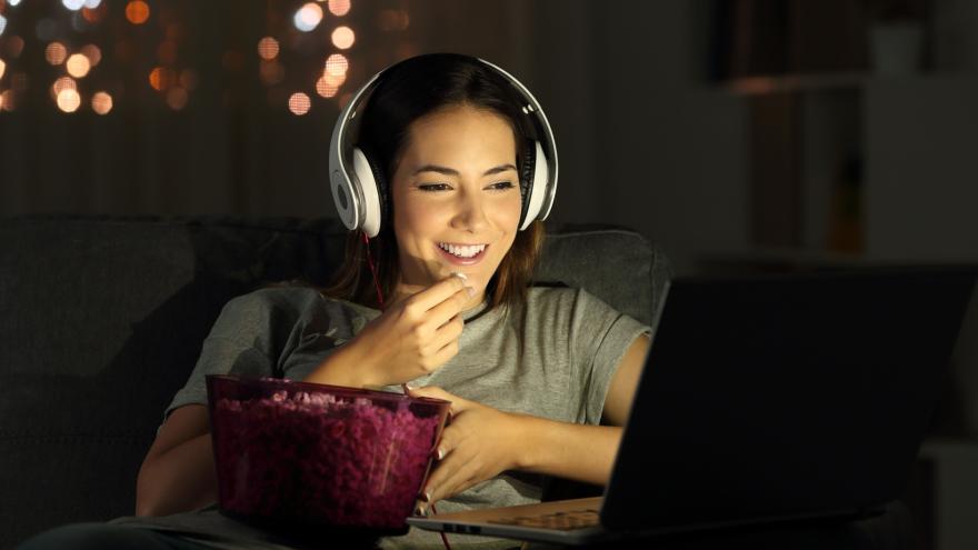 Eine lächelnde Frau mit weißen Kopfhörern und einer Schale Popcorn , die nachts im Internet Fernsehen schaut.