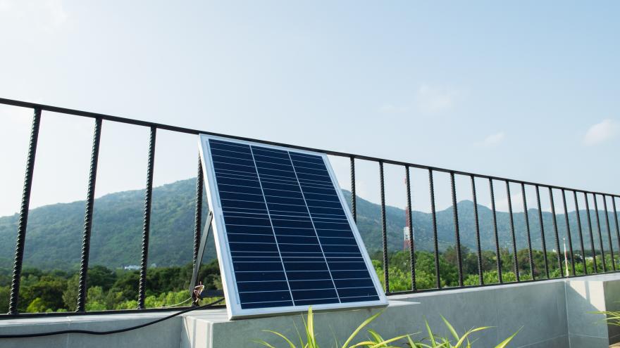 Auf einer sonnigen Dachterrasse lehnt ein einzelnes Solarmodul leicht schräg an einem schwarzen Metallgeländer. Im Hintergrund sind bewaldete Hügel und ein klarer Himmel zu sehen. Das Panel ist über ein Kabel angeschlossen, das über die niedrige Mauer der Terrasse führt. Vorne im Bild ragen einige grüne Zierpflanzen ins Bild und setzen einen natürlichen Akzent zu der technischen Anlage.