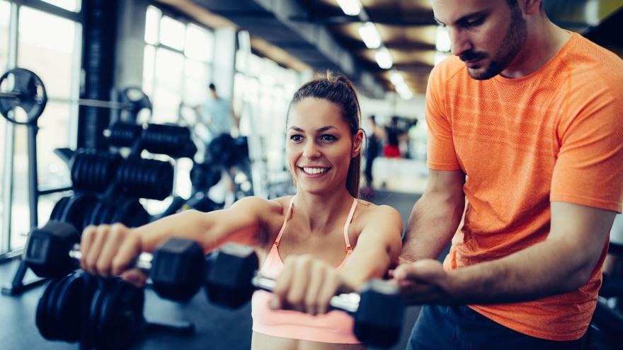 Frau trainiert mit Hanteln im Fitnessstudio, ein Trainer unterstützt sie dabei.