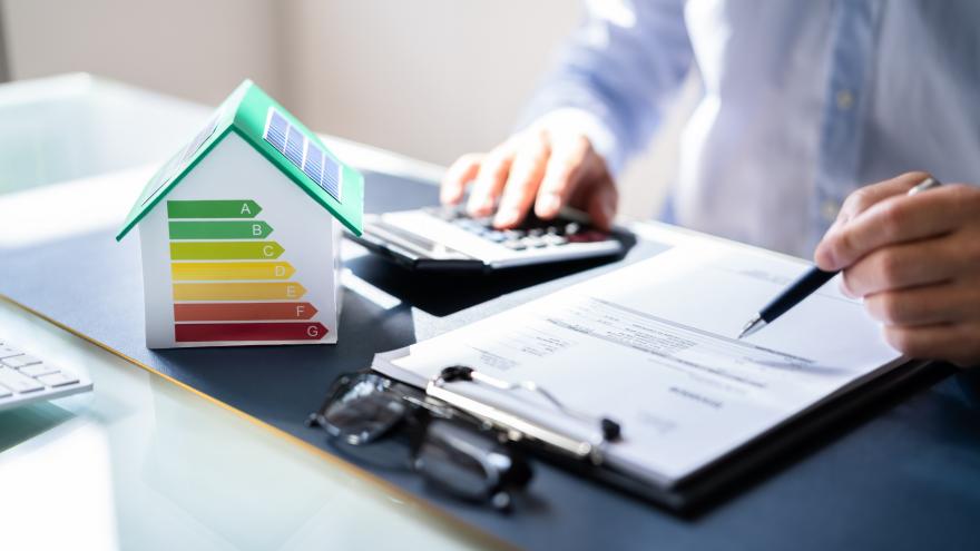 Modellhaus mit Energieeffizienzskala auf einem Schreibtisch neben Unterlagen und Taschenrechner, eine Person berechnet Energiekosten.