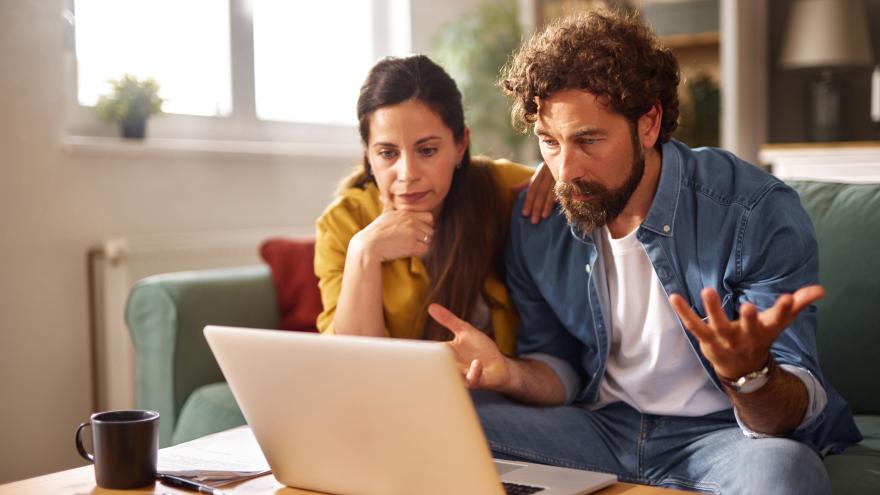 Ein Paar wirkt gestresst, während es auf einer Couch sitzt und auf einen Laptop schaut. Sie sind von Dokumenten und einer Kaffeetasse umgeben, was auf eine ernste Diskussion über finanzielle Probleme hindeutet.