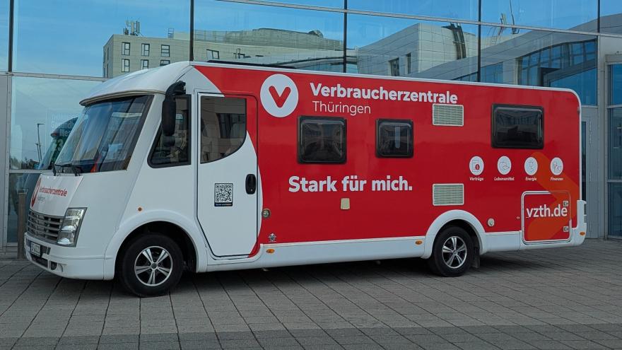 Ein großes, rot-weiß gestaltetes Infomobil der Verbraucherzentrale Thüringen steht vor einem modernen Gebäude mit Glasfassade. Auf der Seite des Fahrzeugs sind das Logo, der Schriftzug ‚Verbraucherzentrale Thüringen‘ sowie der Slogan ‚Stark für mich.‘ zu sehen. Außerdem sind Symbole für Themen wie Verträge, Lebensmittel, Energie und Finanzen abgebildet.