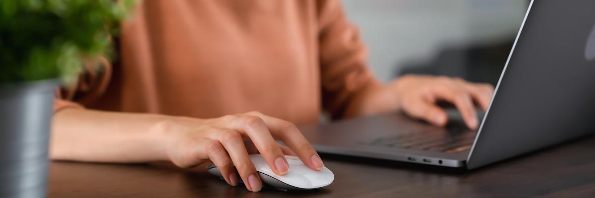 Eine Frau sitzt an ihrem Notebook und hält ihre Hand über eine Computermaus.