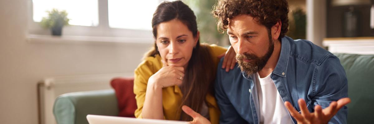 Ein Paar wirkt gestresst, während es auf einer Couch sitzt und auf einen Laptop schaut. Sie sind von Dokumenten und einer Kaffeetasse umgeben, was auf eine ernste Diskussion über finanzielle Probleme hindeutet.