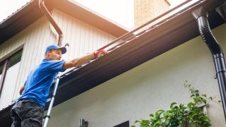 Ein Mann steht auf einer Leiter und reinigt die Dachrinne seines Hauses.
