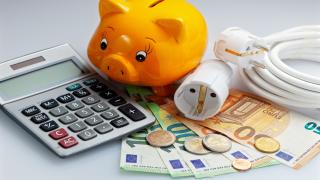 Ein Taschenrechner, ein Sparschwein, Geldscheine und ein Stromkabel sind zu sehen.