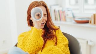 Eine rothaarige Frau in einem gelben Pullover schaut durch gerolltes Papier wie durch ein Fernrohr.