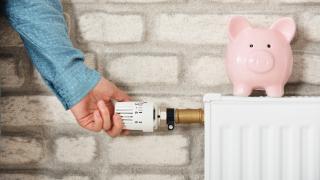 Eine Hand dreht an einem Heizungsthermostat, auf dem Heizkörper steht ein Sparschwein.