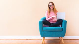 Eine junge Frau sitzt mit einem Laptop in der Hand auf einem blauen Sessel.