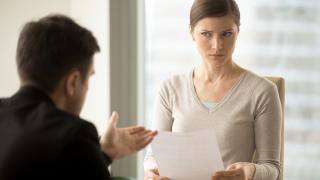 Geschäftsfrau mit skeptischem Gesichtsausdruck, die ein Vertragsdokument in der Hand hält und einem wenig überzeugenden Angebot ihres Geschäftspartners zuhört. 