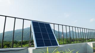 Auf einer sonnigen Dachterrasse lehnt ein einzelnes Solarmodul leicht schräg an einem schwarzen Metallgeländer. Im Hintergrund sind bewaldete Hügel und ein klarer Himmel zu sehen. Das Panel ist über ein Kabel angeschlossen, das über die niedrige Mauer der Terrasse führt. Vorne im Bild ragen einige grüne Zierpflanzen ins Bild und setzen einen natürlichen Akzent zu der technischen Anlage.