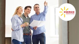 Drei Personen stehen in einem hellen Raum: Ein Mann hält einen Bauplan in der Hand und zeigt nach oben, daneben lächeln ein Mann und eine Frau – rechts ist das Logo „Energieberatung“ zu sehen.