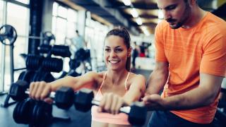 Frau trainiert mit Hanteln im Fitnessstudio, ein Trainer unterstützt sie dabei.